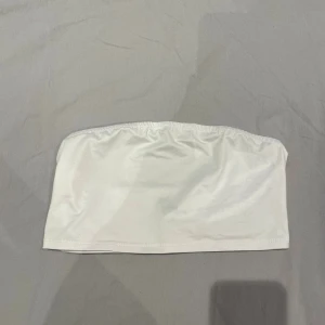 Vit tubtopp - Säljer en enkel och stilren vit tubtopp med elastisk kant upptill och nedtill för att sitta på plats. Toppen är ärmlös och har en slät, mjuk yta i stretchigt material som känns bekvämt mot huden. Perfekt att styla med jeans eller kjol.