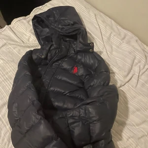 Mörkblå dunjacka från U.S. Polo Assn. - Säljer en mörkblå dunjacka från U.S. Polo Assn. med huva och dragkedja. Jackan har ett broderat rött emblem på bröstet och text på insidan. Perfekt för kyliga och lite varmare dagar. Priset går absolut att diskutera 