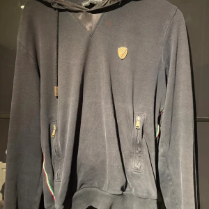 Svart hoodie från Dolce & Gabbana - Svart hoodie från Dolce & Gabbana med huva och guldfärgade dragkedjor på fickorna. Tröjan har ett litet D&G-emblem på bröstet och detaljer med röd, grön och vit rand längs axlarna. Mjuk bomullskänsla och ribbade muddar vid ärmslut och nederkant.