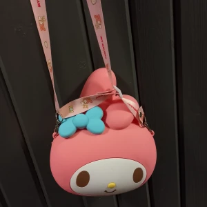 My Melody axelväska i rosa silikon - Supergullig axelväska formad som My Melody från Sanrio. Väskan är rosa med ett stort vitt ansikte, bruna ögon och en blå rosett. Axelremmen är dekorerad med My Melody-figurer och text. Tillverkad i mjukt silikonmaterial och stängs med dragkedja.