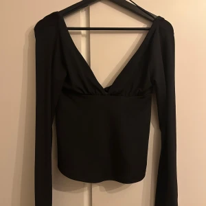 Svart långärmad topp med knyt - Säljer en svart långärmad topp med djup v-ringning och snygg knyt baktill. Toppen är stilren, slät design som passar perfekt till jeans eller kjol. Materialet känns mjukt och stretchigt, vilket gör den bekväm att bära hela dagen.