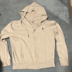 Grå hoodie från Polo Ralph Lauren - Snygg beige hoodie från Polo Ralph Lauren i slim fit-modell. Tröjan har huva med snörning, dragkedja framtill och två fickor. Materialet är mjuk bomull och den klassiska blå Polo-loggan är broderad på bröstet. Perfekt för chill dagar.