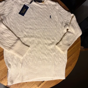 Vit kabelstickad tröja Polo Ralph Lauren - Vit kabelstickad tröja från Polo Ralph Lauren i mjuk ullmix. Klassisk rund hals och ribbade muddar. Diskret broderad logga på bröstet. Perfekt för dig som gillar clean och stilren look med premiumkänsla.