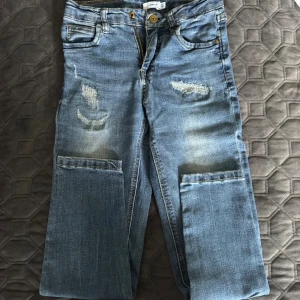 Blå jeans med slitningar från Name It - Snygga blå jeans från Name It med slitna detaljer och stora fickor på benen. Klassisk femficksmodell med dragkedja och knapp. Jeansen har en rak passform och är perfekta för en avslappnad stil.