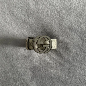 Gucci bälte  - Snyggt Gucci-bälte i beige med klassiskt GG Supreme-mönster och stor silverfärgad GG-logga som spänne. Bältet är gjort i skinn och har en lyxig känsla med detaljerad sömnad och ikonisk design. Perfekt accessoar för att lyfta din outfit.