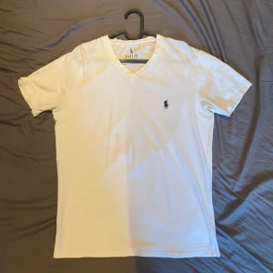 Vit v-ringad t-shirt från Polo Ralph Lauren - Vit t-shirt från Polo Ralph Lauren med v-ringning och klassisk broderad logga i mörkblått på bröstet. Skön passform och tillverkad i mjuk bomull. Perfekt basic-plagg med stilren look.