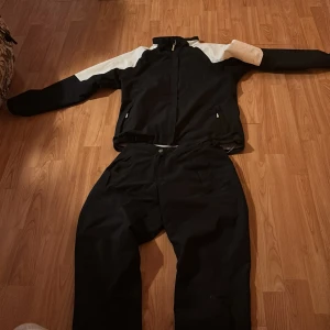 Svart och vit träningsdress från Abacus - Säljer en svart och vit träningsdress från abacus Setet består av en jacka med dragkedja och vita partier på axlarna samt matchande svarta byxor. Perfekt för utomhusaktiviteter och träning. Jackan har fickor med dragkedja och logga på ärmen.