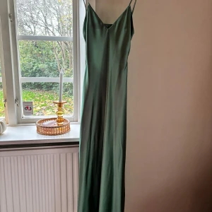 Grön långklänning från Zara - Säljer en elegant grön långklänning från Zara i satinliknande material. Klänningen har smala axelband, v-ringad front och en snygg öppen rygg. Perfekt för dig som gillar minimalistisk och stilren design med lite extra edge. Köpte för 400kr.