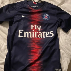 PSG matchtröja Nike blå röd - Paris Saint-Germain fotbollströja i mörkblått med röd grafisk detalj i mitten och vita logotyper. PSG-emblem på bröstet, Fly Emirates-tryck och Nike swoosh. Tillverkad i lätt och ventilerande material, perfekt för match eller träning.
