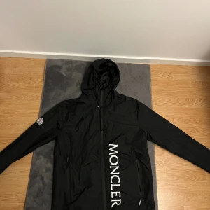Svart vindjacka från Moncler - Svart vindjacka från Moncler med stor vit logotyp längs framsidan och klassisk Moncler-patch på ärmen. Jackan har huva, dragkedja och två sidofickor med zip. Priset kan ändras