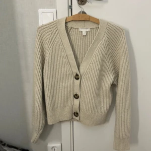 Beige ribbstickad kofta med knappar - Snygg beige stickad kofta med stora bruna knappar framtill. Koftan har v-ringning, lång ärm och breda muddar. Perfekt att slänga över en t-shirt eller linne för en chill look. Mjuk och skön, passar till allt. Plagget är lite knopprig! Pris kan diskuteras🥰