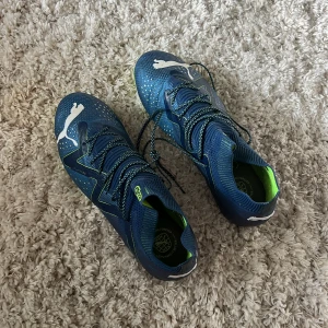  Puma fotbollsskor - Säljer ett par blåa Puma fotbollsskor med coola neon- och limegröna detaljer. Skorna har snörning, mönstrad ovandel och vit Puma-logga. Yttersulan är i neonfärg och har dobbar för bra grepp på planen. Perfekta för dig som vill sticka ut på matchen!