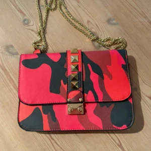Röd och svart camouflage axelväska - Cool axelväska med röd och svart camouflage-mönster, guldfärgade nitar och kedja. Väskan har en rektangulär form och stängs med ett snyggt guldfärgat spänne. Perfekt för dig som vill sticka ut med din stil.