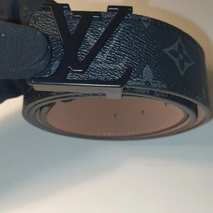 Svart märkeskläder för endast 250 kr. - Snyggt svart märkesbäöte i skinn med klassiskt monogrammönster och stor LV-logga i svart metall som spänne. Bältet är 40 mm brett och har en lyxig känsla med beige insida och fina sömmar. Perfekt accessoar för att lyfta din outfit.