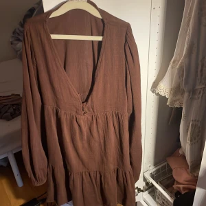 Brun kortklänning från Pull&Bear XS - Superfin brun kortklänning från Pull&Bear i storlek XS. Klänningen har v-ringning med knappar framtill, långa ärmar med resår vid handlederna och ett luftigt, rynkat fall. Perfekt för dig som gillar en avslappnad men ändå trendig look.