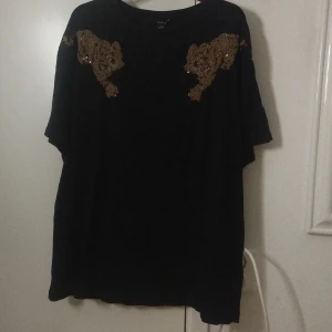 Svart topp med paljett-tigrar från Lindex - Svart loose-fit topp från Lindex med korta ärmar och rund hals. På axlarna finns detaljerade tigrar i guldiga paljetter som ger en cool och unik look. Materialet är mjukt och skönt, perfekt för dig som gillar att sticka ut med djurmotiv och glittriga detaljer.