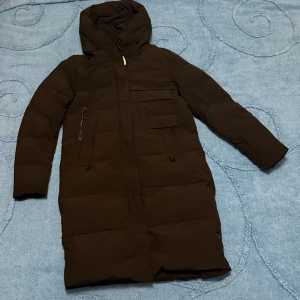 Svart lång pufferjacka  - Svart lång pufferjacka från H-XINZICHENG i storlek XL. Jackan har huva med snörning, flera fickor med dragkedja och knappar framtill. Perfekt för kalla dagar med sin quiltade design och långa ärmar. Snygg och enkel stil som funkar till det mesta.