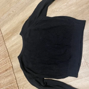 Svart stickad tröja i medium - Svart stickad tröja i storlek medium med ribbade muddar vid hals, ärmslut och nederkant. Tröjan har långärmad design och är gjord i ett grovt stickat material som ger en schysst struktur. Perfekt för att hålla värmen under kyliga dagar.