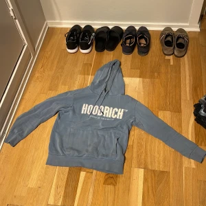 Blå HOODRICH hoodie - Blå hoodie från HOODRICH med vit logga på bröstet. Klassisk känguruficka framtill, huva med snörning och långa ärmar. Skön och avslappnad passform, Tillverkad i mjuk bomullsmix.