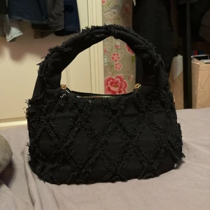 Svart axelväska med quiltad look - Snygg svart axelväska med grovt quiltad yta och fransiga detaljer. Väskan har ett brett handtag och guldfärgade dragkedjor. Medföljer en matchande liten svart plånbok med knappstängning och guldfärgad krok. Perfekt för att lyfta din outfit.