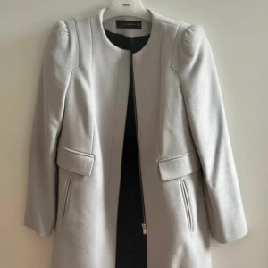 Ljusgrå kappa från Zara - Snygg ljusgrå kappa från Zara Woman med stilren design och rund halsringning. Jackan har långa ärmar, diskreta axelpartier och praktiska fickor med lock. Stängs med dold dragkedja framtill och är fodrad med svart tyg