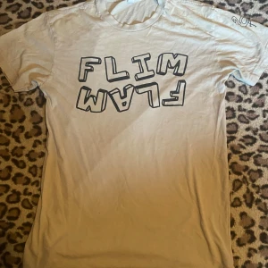 Vit FLIM FLAM t-shirt - Vit t-shirt från Next Level i storlek XS med coolt FLIM FLAM-tryck på bröstet och en flamingo på ärmen. Skön passform och tillverkad i mjuk bomull.