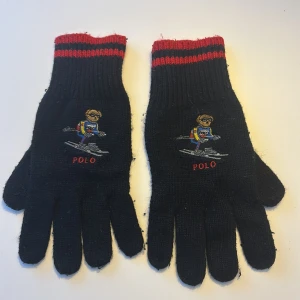Svarta stickade handskar Polo Ralph Lauren - Svarta stickade handskar från Polo Ralph Lauren med röda ränder vid mudden. På ovansidan finns en broderad skidande björn i färgglada kläder och texten POLO. Klassisk design med fem fingrar och mjukt material som håller dig varm.