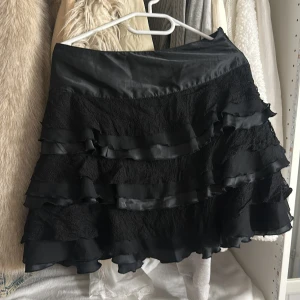 Svart volangkjol från Vila - Supersnygg svart kortkjol från Vila med flera lager volanger och spetsdetaljer. Kjolen har en glansig finish och stängs med dragkedja i sidan. Perfekt för dig som vill ha en kjol med lite extra attityd och rörelse.