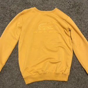 Gul sweatshirt från Lacoste - Gul sweatshirt från Lacoste med stort krokodillogo och text på bröstet. Tröjan har rund hals, ribbade muddar vid ärmslut och nederkant samt långärmad passform. Materialet känns mjukt och skönt, perfekt för chill dagar. Snyggt tryck med flera små krokodiler under loggan.