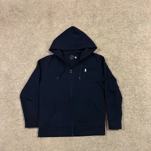 Marinblå hoodie från Polo Ralph Lauren - Snygg marinblå hoodie från Polo Ralph Lauren med dragkedja och huva. Tröjan har broderad logga på bröstet, snörning vid huvan och två praktiska fickor framtill. Perfekt för chill dagar och enkel att matcha med jeans eller joggers.