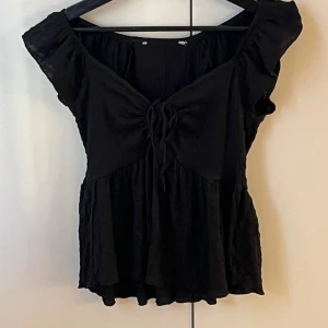Svart blus med knyt och volang - Svart blus med korta ärmar, v-ringning och knyt framtill. Toppen har volangig nederdel och lätt rynkad struktur som ger en luftig känsla. Perfekt för dig som gillar en feminin och trendig stil.