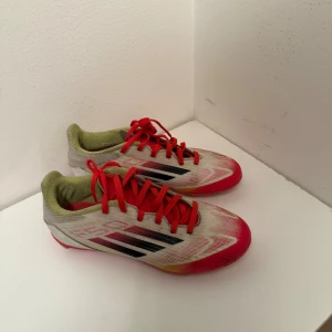 Adidas fotbollsskor - Säljer ett par Adidas fotbollsskor med snygg design i vitt, rött och inslag av gult. Skorna har röda snören, svarta Adidas-ränder på sidorna och röda dobbar undertill. Ovandelen är i syntetmaterial och insidan är ljusgrön. Perfekta för dig som vill sticka ut på planen.