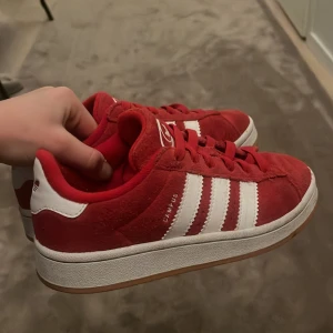 Röda Adidas Campus sneakers - Säljer ett par röda Adidas Campus sneakers i mocka med klassiska vita ränder på sidorna och vit logga på plösen. Skorna har snörning och en platt sula i gummi. Perfekta för dig som gillar retrostil och vill sticka ut med färg.