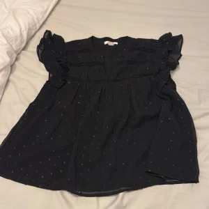 Svart blus med volang och prickar - Säljer en svart blus med glittriga små prickar och volangdetaljer vid axlarna. Blusen har en v-ringad hals och är kortärmad med lätt transparent material som ger en luftig känsla. Passformen är loose och toppen är perfekt för dig som gillar en söt och trendig look.