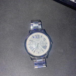 Silverfärgad klocka från Regal - Stilren klocka från Regal med rund urtavla i silver och romerska siffror. Armbandet är i blankt metallmaterial och har en klassisk länkdesign. Urtavlan har flera små visare och detaljer som ger en modern vibe. Perfekt för dig som gillar snygga accessoarer.