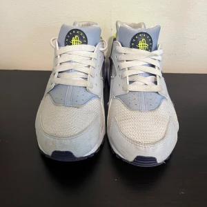 Nike air Huarache - Klassiska och bekväma Nike Air Huarache i stilren vit/grå färg med mörkblå och neon-gula detaljer. En retromodell med en modern twist – perfekt för både vardagsoutfits och streetwear.