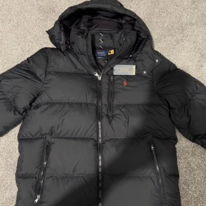 Ralph Lauren Puffer Jacka - Snygg och varm pufferjacka från Ralph Lauren, perfekt för både vardag och stadsliv. Jackan har en klassisk svart färg som passar till allt, med Ralph Laurens ikoniska logga broderad på bröstet. Den är lätt men värmande, fylld med högkvalitativt material som håller dig varm under kalla dagar utan att kännas tung.  ✅ Stilren design med sportig touch ✅ Mjuk och bekväm passform ✅ Praktiska fickor med dragkedja ✅ Elastiska ärmslut och justerbar fåll för perfekt passform
