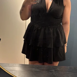 Svart kort volangklänning med knyt - Supersnygg svart kort klänning med djup v-ringning och volanger i kjolen. Smockad midja för skön passform och knyt i nacken som ger en cool halterneck-look. Perfekt för dig som gillar att sticka ut med en feminin och trendig stil.