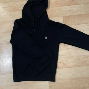 Svart hoodie från Polo Ralph Lauren - Svart hoodie från Polo Ralph Lauren med klassisk vit broderad logga på bröstet. Tröjan har huva med snörning och känguruficka framtill. Skön passform och tillverkad i mjuk bomull, perfekt för chill dagar eller när du vill ha en clean look.