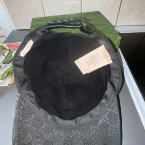 Svart Gucci keps med GG-mönster - Snygg svart keps från Gucci med klassiskt GG-monogram över hela kepsen. Har en grön och röd rand på sidan och justerbar rem baktill i skinn. Materialet är bomull och kepsen har en böjd skärm. Perfekt för dig som vill ha en lyxig streetstyle-look.