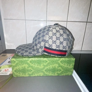 Gucci keps med GG-mönster och röd/blå detalj - Snygg keps från Gucci med klassiskt GG-mönster i grått och blått. Kepsen har en böjd skärm och en ikonisk röd/blå rand på sidan. Justerbar rem baktill för perfekt passform. Materialet är bomull och polyester, perfekt för dig som vill sticka ut med lyxig streetstyle.