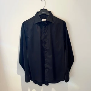 Svart skjorta från Stenströms - Stilren svart skjorta från Stenströms i fitted body-modell. Skjortan har klassisk krage, knappar framtill och långa ärmar med breda manschetter. Materialet är mjukt och är i toppklass bomull, perfekt för dig som gillar en clean look. Hör av er vid frågor