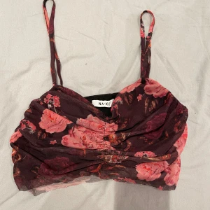 Blommig croppad linnetopp från NA-KD - Supersnygg croppad linnetopp från NA-KD med blommigt mönster i rosa och vinröda toner. Tunn mesh överdel och smala axelband ger en trendig vibe. Perfekt för dig som vill sticka ut med färg och mönster. Säljes då jag rensar garderoben!