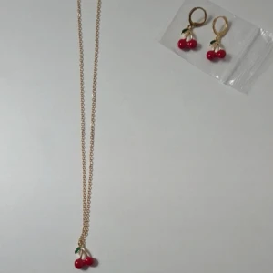 Set med körsbärshalsband och örhängen, 45 cm - Supergulligt smyckeset med ett halsband och ett par örhängen i guldton med körsbärsberlocker. Körsbären är klarröda med gröna blad och ger en lekfull vibe. Perfekt för dig som gillar färgglada detaljer och vill sticka ut lite extra. Halsband: Material: Legering, 18K guldpläterad Längd: 45 cm Hänge: Dubbla körsbär i röd emalj     Örhängen: Modell: Små hoops/creoler med körsbärshänge Material: Kopparlegering Mått (ungefärliga): Total höjd 3 cm Körsbärshänge bredd ca 1.5 cm Skick: Nytt