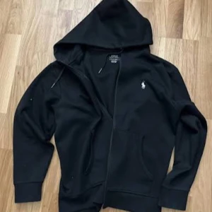 Svart hoodie från Ralph Lauren - Svart hoodie från Ralph Lauren med klassisk vit logga på bröstet. Tröjan har huva med snörning, dragkedja framtill och två fickor. Perfekt för chill dagar och enkel att matcha med jeans eller joggers.