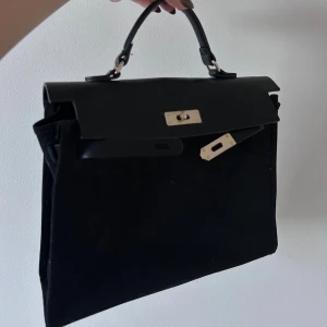 Svart handväska med gulddetaljer - Stilren svart handväska med klassisk topphandtag och snygga guldfärgade metalldetaljer. Väskan har en strukturerad, fyrkantig form och är tillverkad i en kombination av skinn och mocka. Perfekt för dig som gillar minimalistisk design med lyxig känsla. Knappt använd