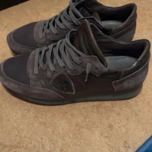 Philippe Model grå sneakers herr - Snygga grå sneakers från Philippe Model i storlek 42. Skorna har en mix av mocka och textil, med klassisk snörning och rund tå. Stilren design med diskret logga på sidan och platt sula, perfekta för en clean och urban look.