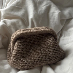 Virkad beige handväska med mönstrad insida - Säljer en handgjord virkad handväska i brunt. Väskan har en mjuk, chunky struktur och är fodrad med ett ljust tyg med grafiskt mönster. Perfekt storlek för det viktigaste och har en unik, retroinspirerad form. Superfin att ha som accessoar till din outfit.