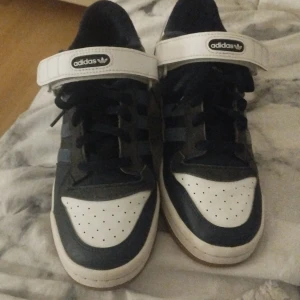 Adidas Forum Mid svart/vit strl 43 1/3 - Säljer ett par Adidas Forum Mid sneakers i mörk blå och vitt med klassiska tre ränder på sidan, snörning och kardborreband upptibll som går att ta bort. Snygg retrostil med gummisula och Adidas-logga på hälen och tungan. Perfekta för dig som gillar streetwear och ikoniska modeller. Kartongen finns kvar till skorna. 