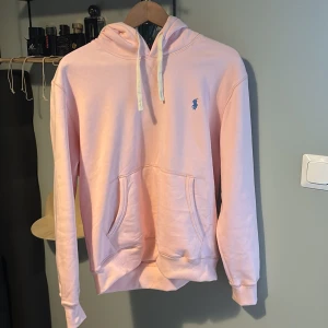 Ljusrosa hoodie från Polo Ralph Lauren - Säljer en ljusrosa hoodie från Polo Ralph Lauren med klassisk huva och dragsnören. Tröjan har en broderad blå logga på bröstet och en stor magficka. Perfekt för dig som gillar stilren och bekväm streetwear.
